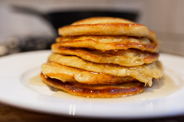 Fresh homemade classic pancakes with maple syrup in a white plate on a wooden board with a pan in the background.