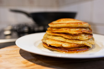 Fresh homemade classic pancakes with maple syrup in a white plate on a wooden board with a pan in the background.