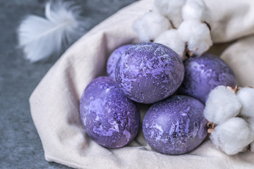 Spotted painted purple eggs of a chicken. Colorful easter eggs. Colored eggs purple on an eco canvas bag with a feather and cotton bolls. Purple eggs on a gray background. Happy easter