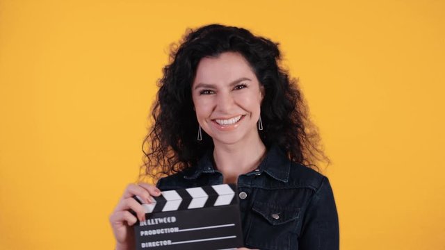 Portrait of beautiful young woman with black curly hair turning into the cam twinking and clapping a clapperboard in front of yellow background shot in 4k