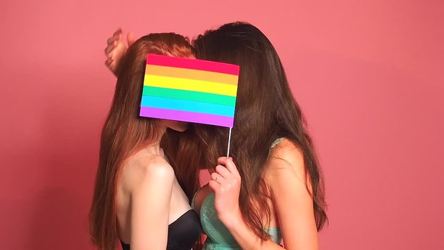redhaired ginger woman in pink studio background kissing and hiding behind the rainbow flag