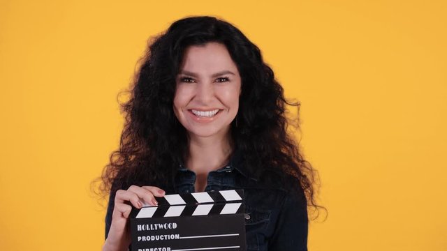 Portrait of beautiful young woman with black curly hair twinking and clapping a clapperboard in front of yellow background shot in 4k