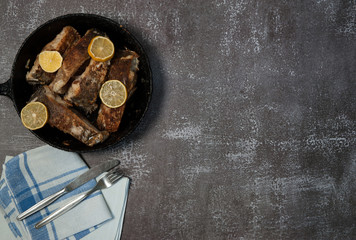 Frying pan with fried pieces of fish bream (Abramis), lemon slices, cutlery and linen napkin on a gray concrete background. Free space.