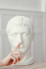 Female hand pokes a finger into the nose of Gypsum sculpture of Caesar's head. Head stands on a wooden table in a classic interior