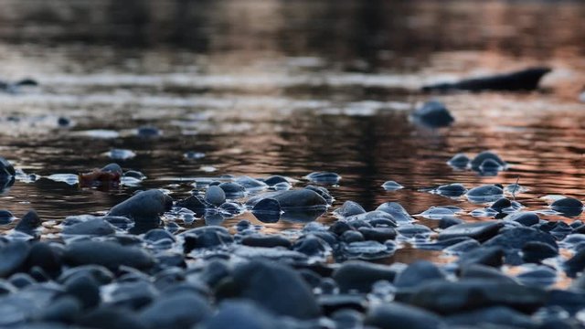 Low vantage loop ready shot of the spring sunset on a rippling river, crimson reflection on the water current with blurred pebbles soft focus.