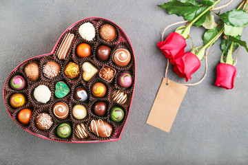 Heart-shaped box with delicious candies and bouquet of flowers on grey background
