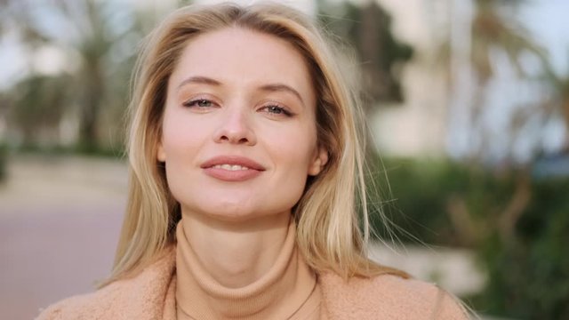 Tracking out shot of charming blond girl happily looking in camera on city street