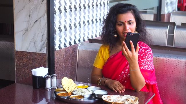 beautiful woman eating dosa chutney naan and indian tali and talking by phone in a restaurant.She wearing red sari with gold earrings , with tilaka on her forehead