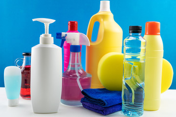 Plastic bottles of cleaning products set with pile clothes on white table blue background.