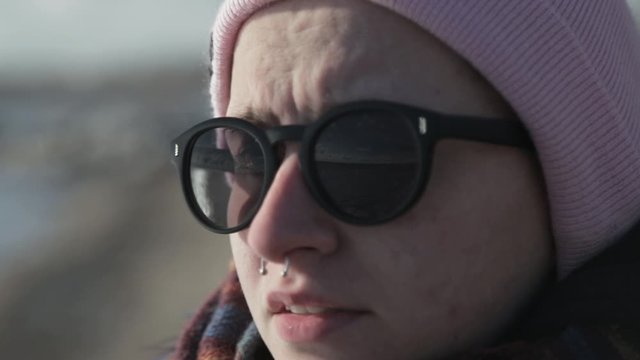 Gorgeous Caucasian Lady Wearing Sunglasses And With A Septum Piercing Staring Somewhere By The Lake Shore In Canada On A Sunny Day - Closeup Shot