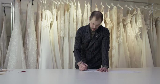 Handsome wedding designer draws dress sketch on a desk in atelier
