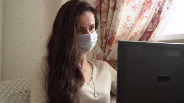 Beautiful woman with long brown hair in protection face mask in beige sweater sit in armchair, works remote on laptop at home close up. Caucasian girl typing on black notebook keyboard in isolation.