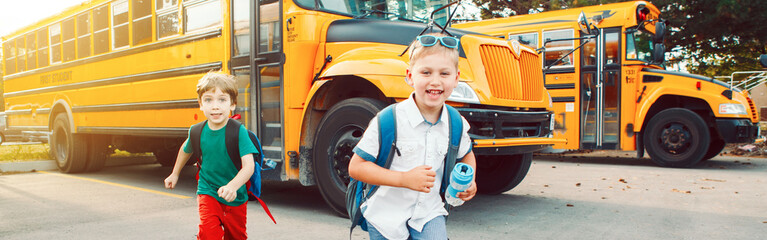 Two funny happy Caucasian boys students kids running near yellow bus on 1 September day. Education back to school. Children ready to learn, study. Web banner header for a website.