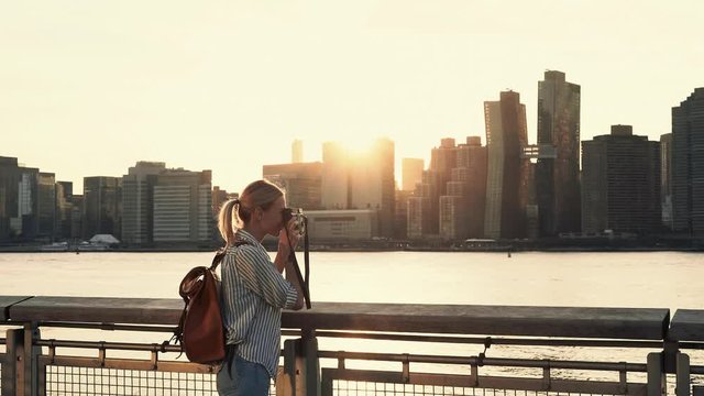 Hipster girl photographing scenic landmark of Manhattan island via film instant camera looking at made image and clicking photo with smartphone application for sharing to travel website
