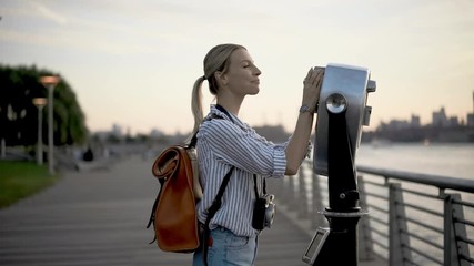 Happy female traveller with touristic backpack using public binocular for looking around metropolitan skyline buildings in New York, smiling hipster girl enjoying getaway for discovering
