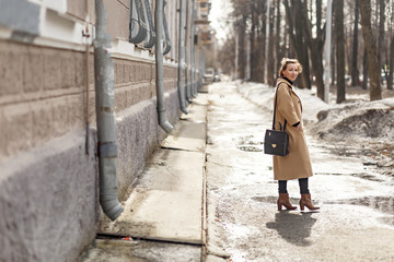 young beautiful girl in a beige coat, dressed in a hipster style, enjoys the spring weather and the sun, walks around the city one spring morning