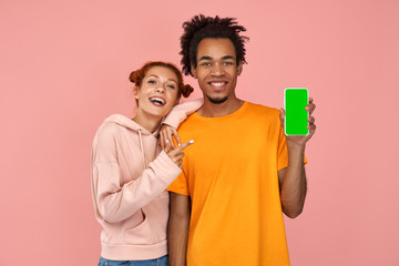 Selective focus of happy ginger woman and dark skinned man points to modern device with green screen with alfa channel for your advertising, promote cool gadget, have smiles, stands over pink wall