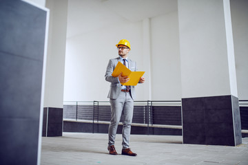 Full length of handsome classy unshaven male architect in suit and with helmet on head walking outdoors with folder in hands.
