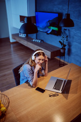 Fototapeta na wymiar Top view of lovely Caucasian young brunette sitting at dining table and listening music over headphones. On table is laptop.
