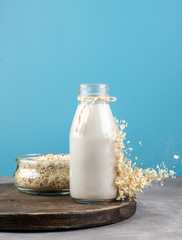 Oat Milk in a bottle with splash of oat flakes and oat flakes in a jar on blue background.