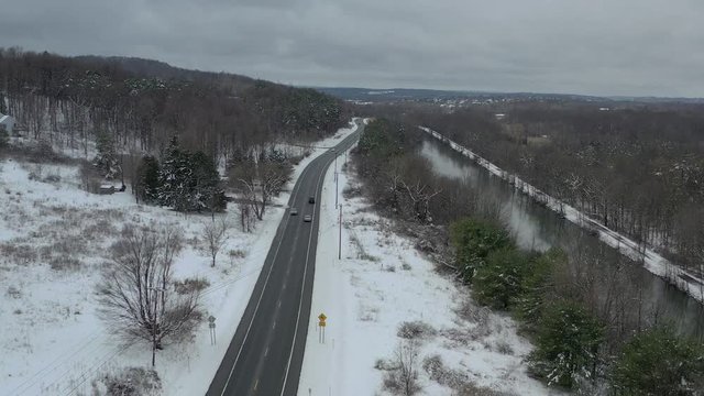 Flying over trees as multiple vehicles travel along highway next to river in winter on overcast day.