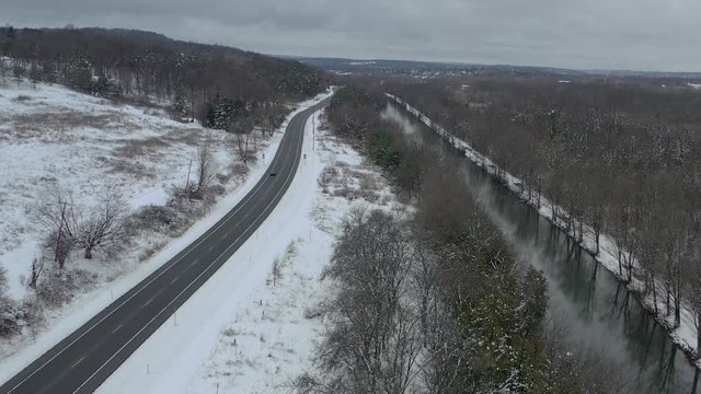 Single black car traveling down highway next to river in winter on overcast day. Distant aerial, dolly in