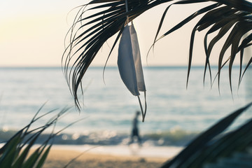 Hygienic mask hanging on palm leaves on the beach against the sea