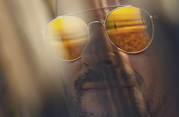 Close-up portrait of a man's face in orange sunglasses, which reflects the beach, the sea and the...