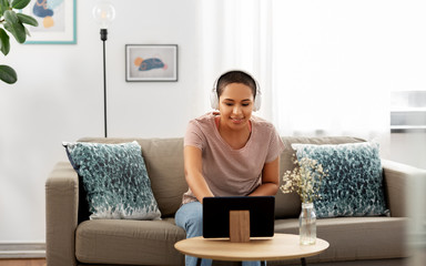 people, technology and leisure concept - happy smiling young african american woman in headphones sitting on sofa and listening to music or watching movie on tablet pc computer at home