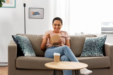 Obraz premium people and leisure concept - happy smiling african american young woman eating takeaway food with chopsticks at home