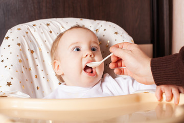 The child is funny, sitting on a highchair, scared. A one-year-old girl in a white sweater is sitting at a yellow table on a brown background, waiting for food with frightened big eyes.