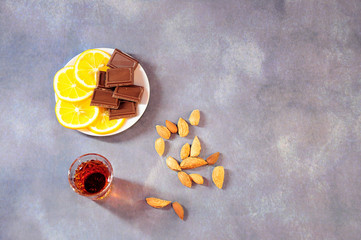 A glass of cognac and a plate with slices of lemon and chocolates, next to a scattering of almonds.