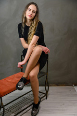 Vertical studio portrait of a caucasian young pretty girl in a black dress with long hair sitting on a stand against a gray background.