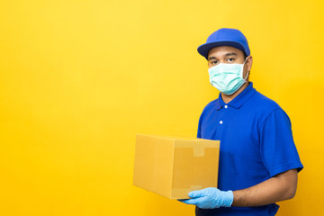 Delivery man blue uniform wearing rubber gloves and mask holding parcel cardboard box on yellow background.