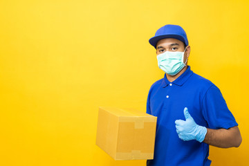 Fototapeta na wymiar Delivery man blue uniform wearing rubber gloves and mask holding parcel cardboard box on yellow background.