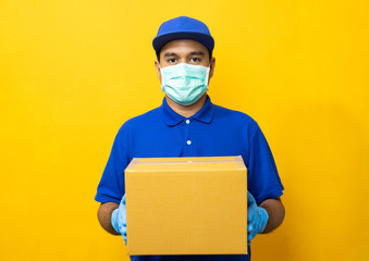 Delivery man blue uniform wearing rubber gloves and mask holding parcel cardboard box on yellow...
