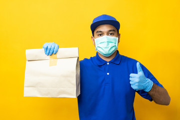 Fototapeta na wymiar Delivery man blue uniform wearing rubber gloves and mask holding paper bag on yellow background.