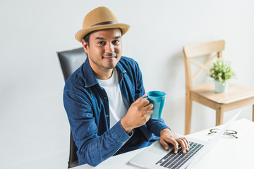 Work from home of young asian man holding and drinking a cup of coffee in the morning. Casual asian businessman age around 30 using on laptop for his work.