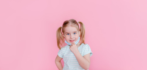 Optimistic, smiling happy little girl taking off the medical mask from face on a pink background showing end of the pandemia. Copyspace.