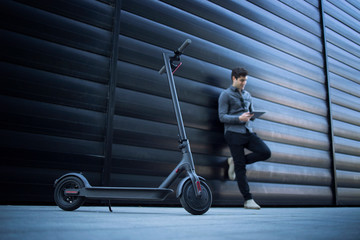 Electric scooter. Eco friendly modern way of transportation. In background handsome business person caucasian man using tablet computer leaning on black business building. © littlewolf1989
