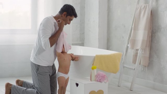 Medium shot of caring Indian father in casual clothes undressing cute toddler girl and putting her into bathtub