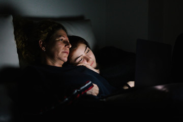 Fototapeta premium Mother and daughter on the couch watching a movie on the computer with the lights off