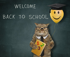 The beige cat in a black tie and glasses is holding a yellow smiling balloon in a square academic cap and a textbook near a blackboard with lettering welcome back to school.
