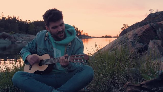Young Musician playing guitar and singing in front of bonfire at the ocean. Guy is singing in front of friends during sunset while traveling.