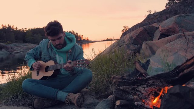 Young man singing with acoustic guitar in front bonfire and sea on beautiful lonely island.