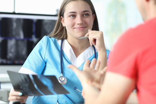 Close-up of smart young doctor discussing x ray results with patient. Smiling specialist holding scan of hand. Modern medicine and professional treatment concept