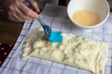 Female hands brushing beaten egg with a brush on the surface of a raw strudel.