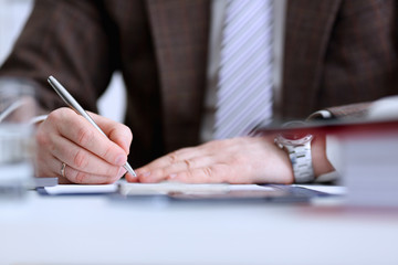 Male arm in suit and tie hold silver pen filling schedule in notepad at office workplace closeup....