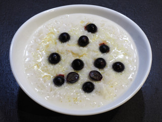 Milk rice porridge with butter and blue berries in a white bowl closeup on a dark background, top view. Healthy grain pudding with cream for children for breakfast
