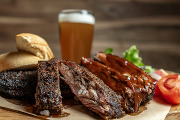 Smoked pork ribs with tomatoes, salad and bread on wood cutting board with glass of beer on background
